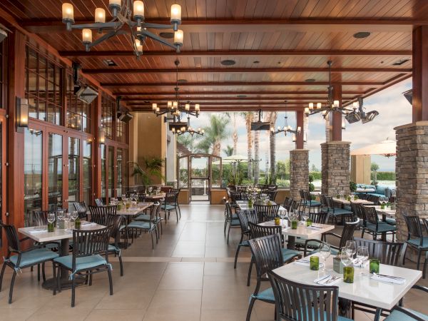 A stylish restaurant patio with long dining tables, hanging lantern lights, a wood ceiling, and stone columns, ready for guests.