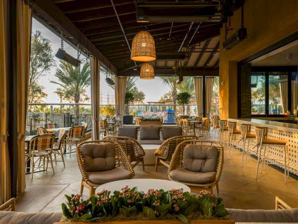 A warm, covered outdoor patio with rattan lounge chairs, hanging pendant lights, and a view of trees at sunset.