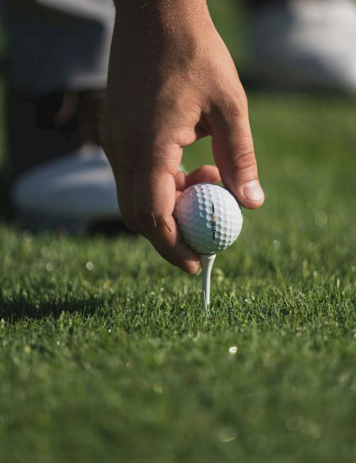 A close-up of a golf ball on a tee, about to be struck, with a golfer&rsquo;s hand guiding it on a grassy tee box.