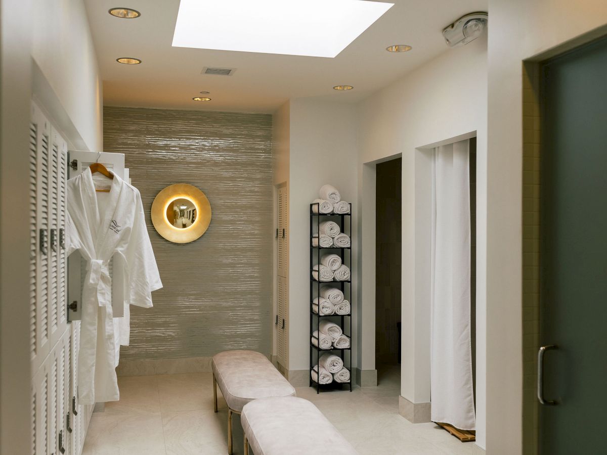A hotel spa hallway shows white bathrobes, a bench, rolled towels, a round lit mirror, and soft ceiling lights.