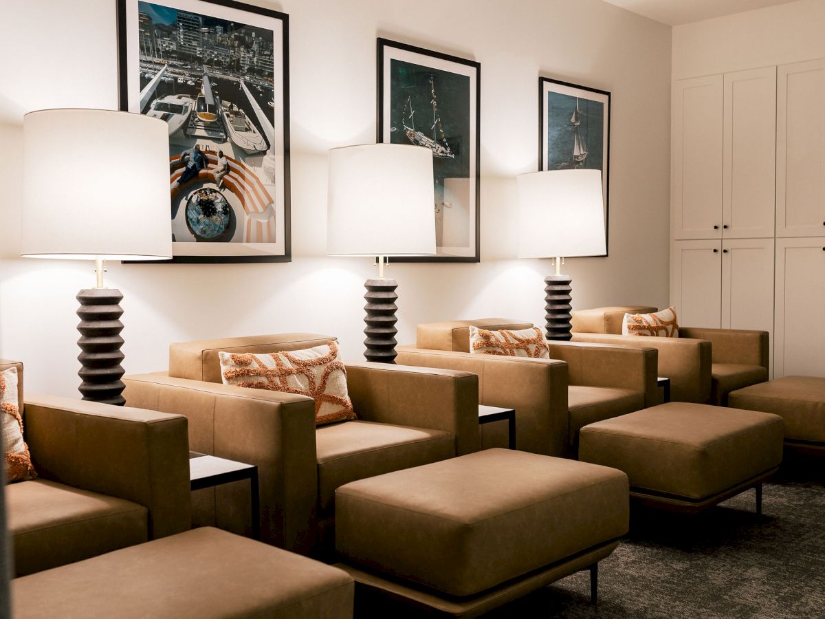 A modern lounge with tan armchairs, matching ottomans, tall stacked lamps, framed wall art, and a bright, calming seating area. The scene is cozy and contemporary.