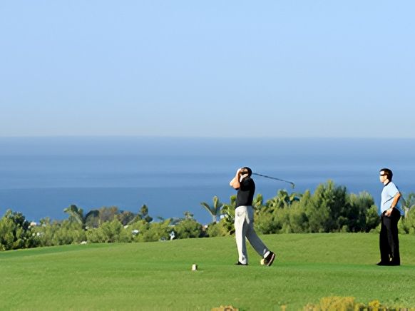 A golfer is swinging on a sunny hilltop golf course with the ocean in the background, while a man watches nearby.