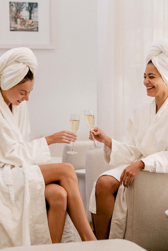 Two women in robes with towels on their heads toast glasses of champagne in a spa/lounge setting, smiling and relaxing together. The line ends with a period.