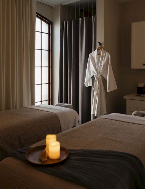 A cozy spa-like hotel room with two neatly made beds, a wall of windows, a robe on a hanger, and lit candles on a tray by the bed.