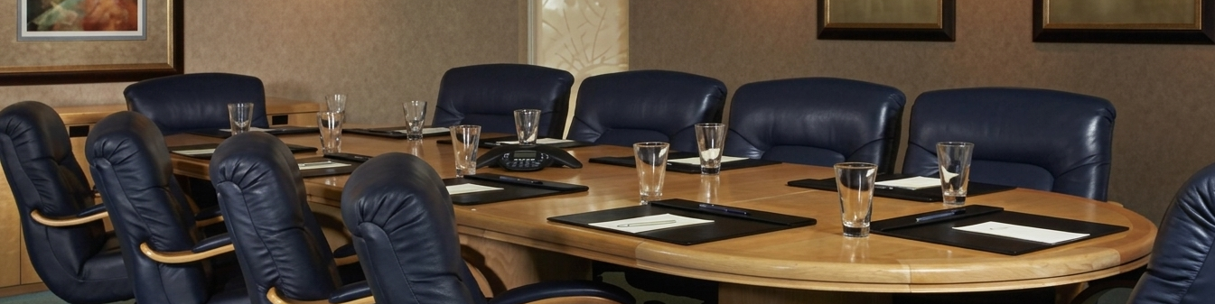 A boardroom conference table with navy leather chairs, glasses of water, phones, and framed art on brown walls.