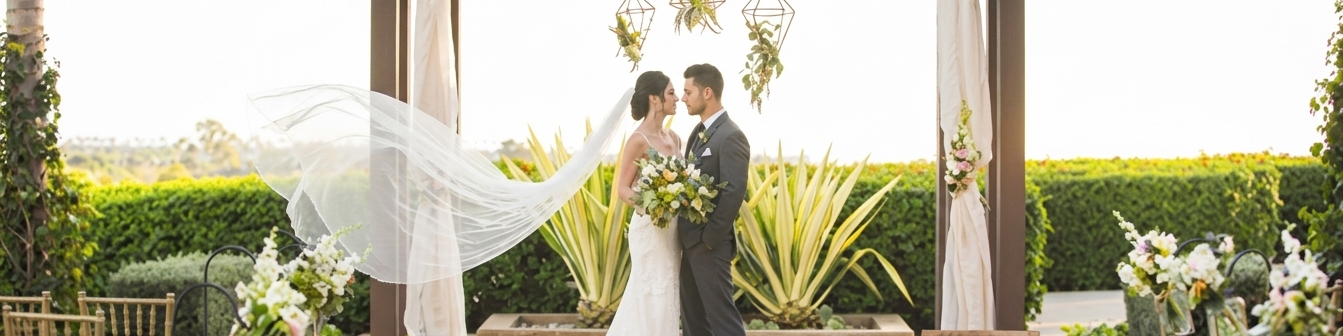 An outdoor wedding ceremony setup with a bride and groom under a floral draped arch, surrounded by chairs and greenery.