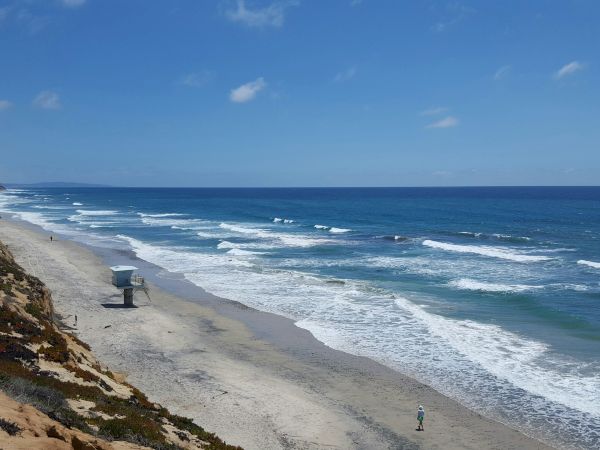A sunny coastline with rolling waves, a sandy beach, cliffs, and a lifeguard stand under a bright blue sky.