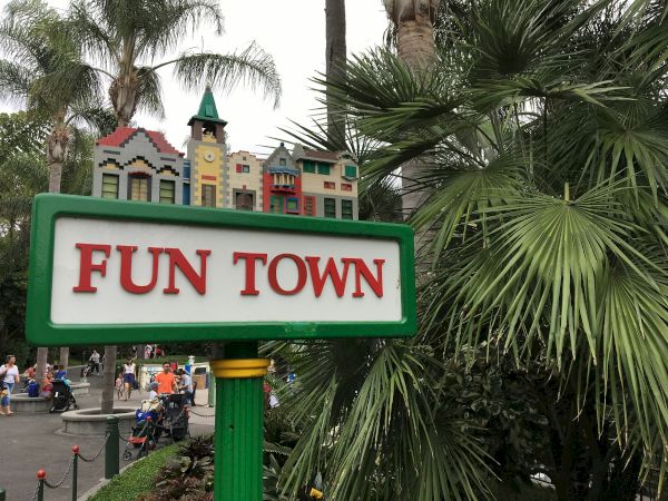A colorful &ldquo;Fun Town&rdquo; sign with playful buildings on top, tropical palm trees, and people in a theme-park setting.