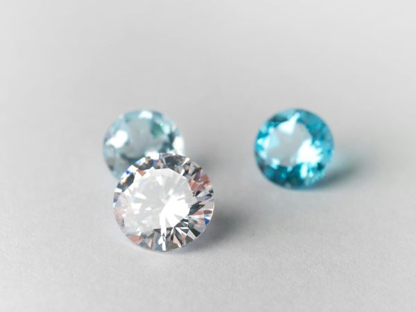 Three sparkly gemstones on a pale surface: a large clear diamond, plus two smaller blue stones, one darker teal, one lighter aqua.