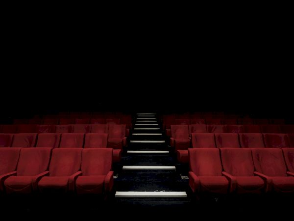 Empty red theater seats with a single lit center aisle stairs fading into darkness, suggesting a silent, awaiting performance.