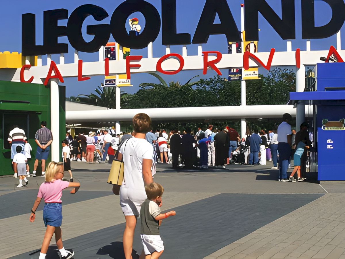 A sunny Legoland theme park entrance with a huge &ldquo;LEGOLAND CALIFORNIA&rdquo; sign and families walking toward attractions.