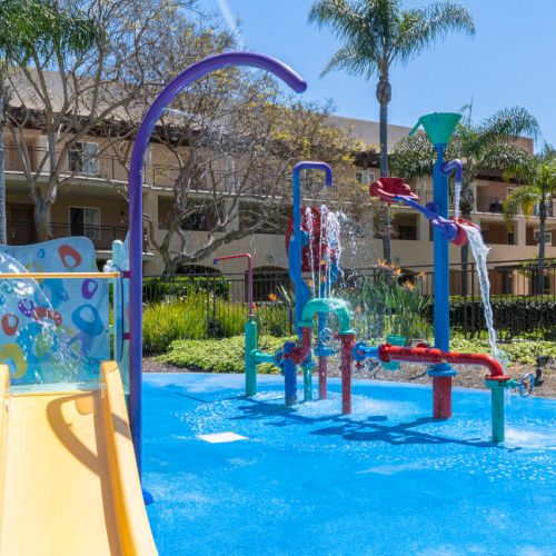 A sunny apartment splash pad with bright water slides, fountains, and spraying jets in a fenced pool area.