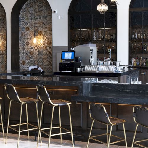 A stylish caf&eacute; bar with ornate tile arches, stocked liquor bottles, espresso/cocktail equipment, and tall stools in front.