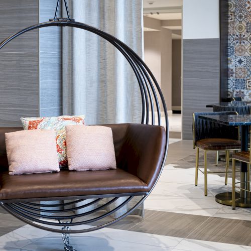 A modern indoor lounge with a suspended round hanging chair, cushioned pillows, and a patterned wall with a small dining area.