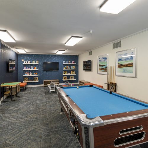 An indoor game room with a blue billiards/pool table, cushioned seating, shelves with decor, and colorful wall art.