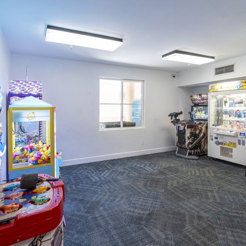 An indoor arcade room with prize machines, ball/ticket games, bright lights, and colorful claw-style arcade cabinets.