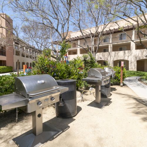 A sunny apartment courtyard with stainless BBQ grills, a trash can, shrubs, and leafless trees beside beige buildings.