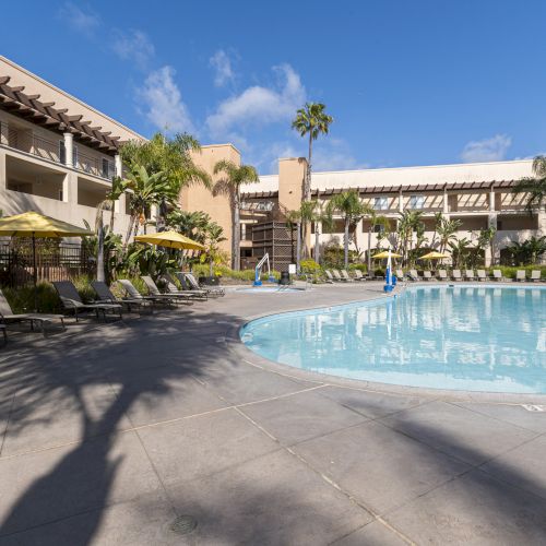Sunny hotel courtyard with a clear swimming pool, loungers, palm trees, and balconies under a blue sky.