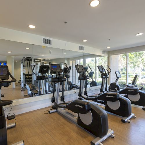 A bright fitness room filled with treadmills and stationary exercise bikes near large windows and a wall of equipment.