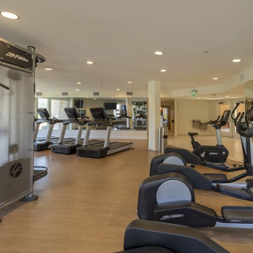A bright gym room with treadmills and cardio equipment lined along the walls under ceiling lights.