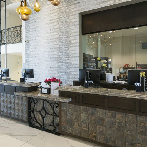A modern hotel reception with a patterned marble front desk, glass windows, and hanging amber chandeliers in a bright lobby.
