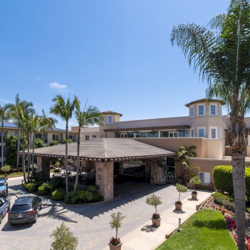 A sunny hotel courtyard with palm trees, manicured gardens, parked cars, and beige buildings under a clear blue sky.