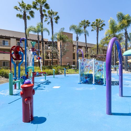 A sunny splash playground with colorful water sprayers in a pool, surrounded by palm trees and apartment buildings, all gleaming.