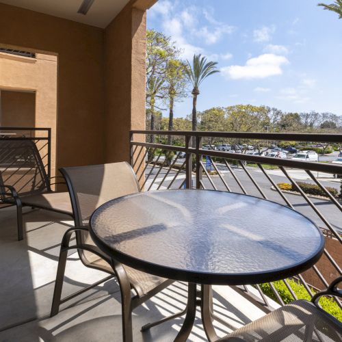 A sunny apartment balcony with a round patio table and chairs overlooking cars, trees, and tall palm trees.