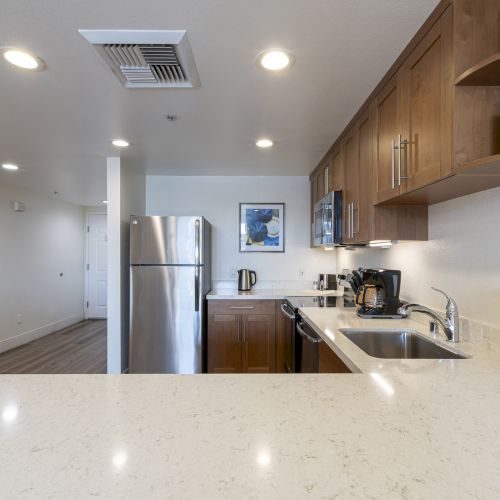 A bright modern kitchen with wood cabinets, stainless fridge, granite counters, double sink, coffee maker, and recessed ceiling lights.