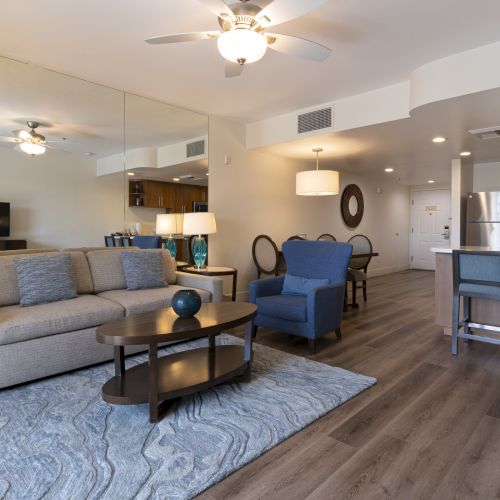 Bright modern living room with sofas, coffee table, kitchen bar, stainless fridge, ceiling lights, and a large TV.