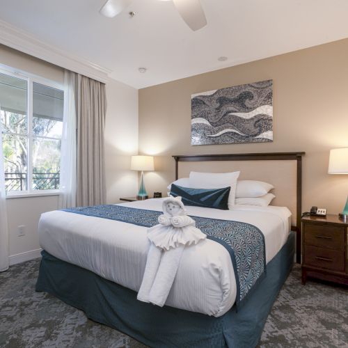 A bright hotel bedroom with a neatly made bed, bedside tables, lamps, large window curtains, and ocean-themed wall art.