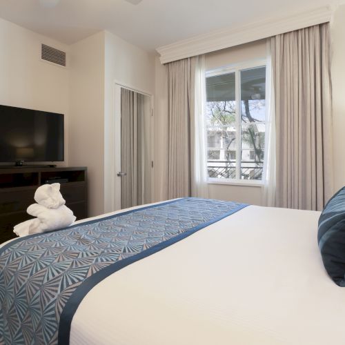 A bright hotel-style bedroom with a large bed, flat-screen TV, and curtains framing a window with daylight.