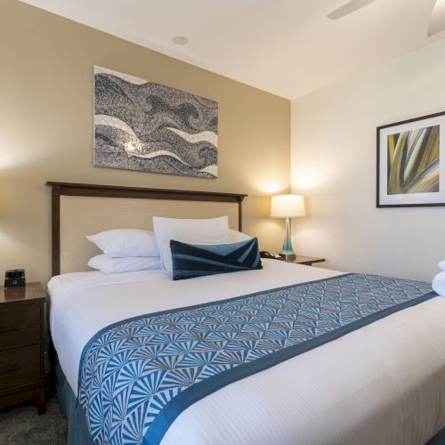 A neatly made hotel bedroom with a bed, two lamps, and wall art featuring abstract ocean waves and plants.