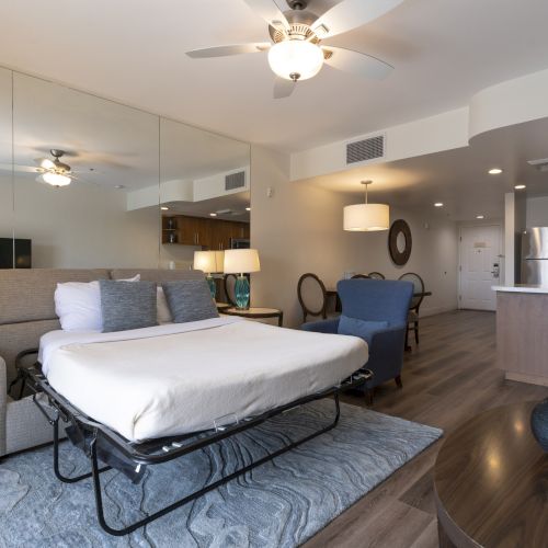 Modern hotel room with a bed, ceiling fan lights, a seating area, and a kitchen bar with stools.