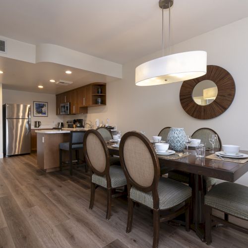 Modern dining room with set table, wood chairs, pendant lamp, and an adjoining kitchen with cabinets and fridge.