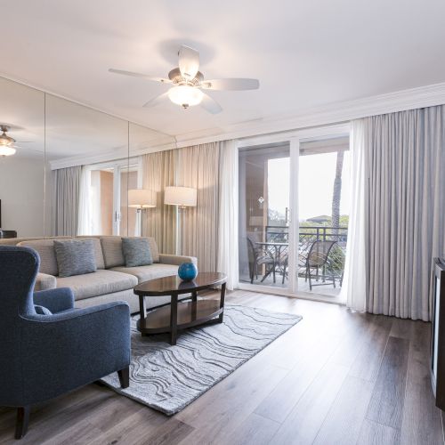 A modern living room with a gray sofa, coffee table, flat-screen TV, floor-to-ceiling curtains, and sliding balcony doors.