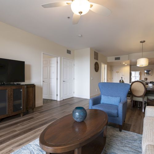 Bright living room with a TV, blue armchair, coffee table, dining area, and kitchen, illuminated by ceiling lights and fans.