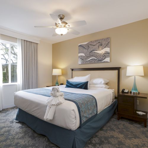 A tidy hotel bedroom with a made bed, bedside lamps, framed wall art, curtains, and a ceiling fan overhead.