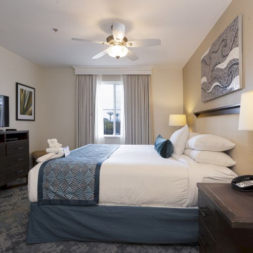 A hotel bedroom with a king-size bed, ceiling fan light, TV, bedside lamps, and large window with curtains.