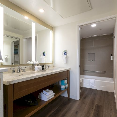 A bright hotel bathroom with a marble vanity, double sinks, mirrors, towels, and a bathtub with grab bar.