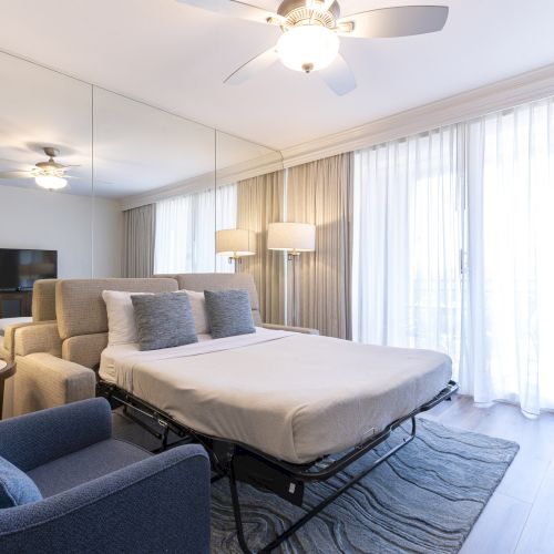 A modern hotel bedroom with a large bed, blue armchairs, floor-to-ceiling curtains, and a lit ceiling fan, in a bright airy room.